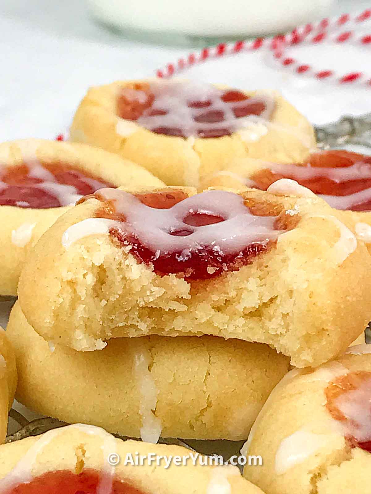 A close shot of a thumbprint cookie filled with jam and topped with glaze and a red and white bakers twist at the background.