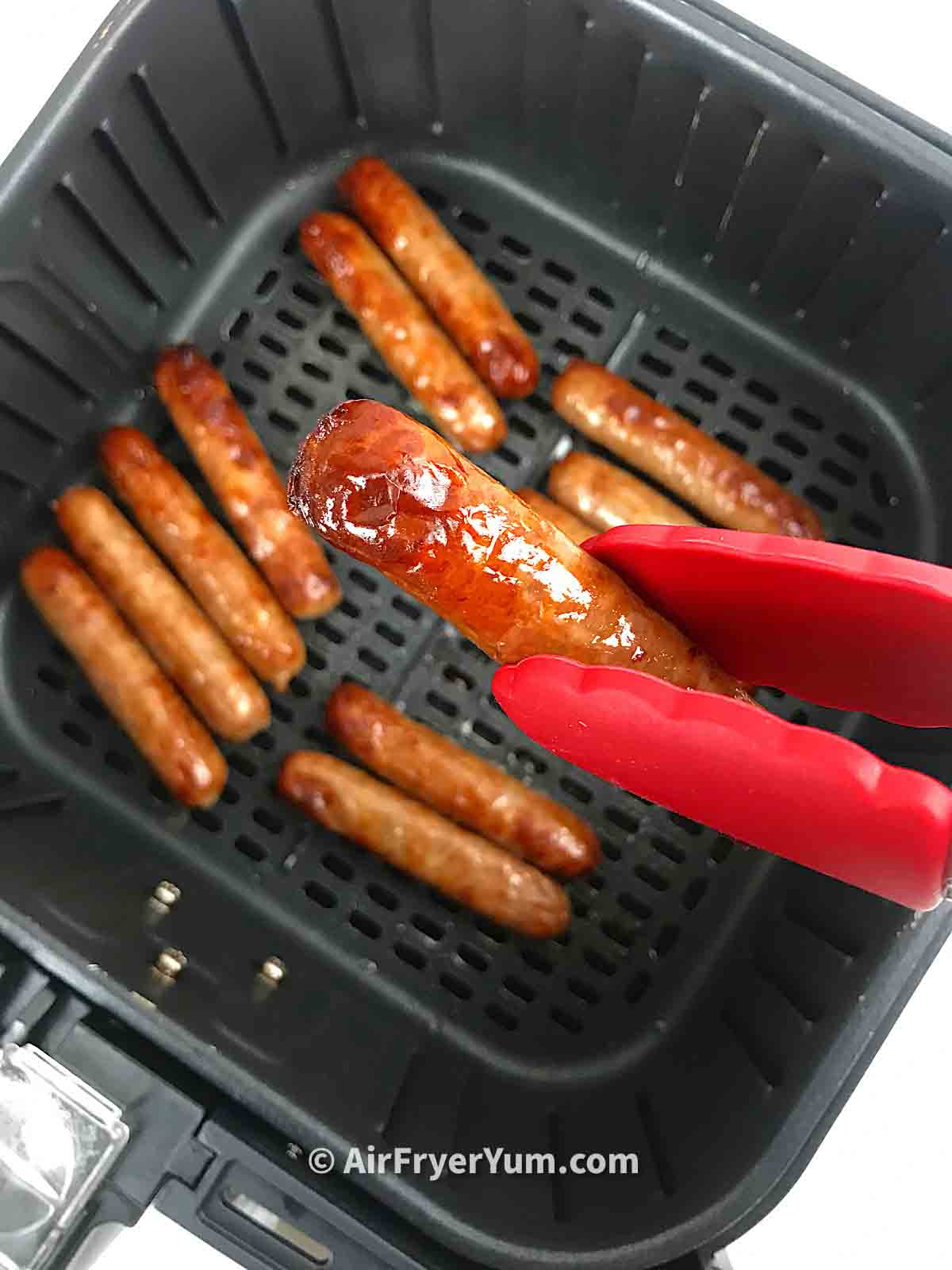 Sausage lifted up with a pair of red tipped kitchen tongs and more sausages in the air fryer basket at the background
