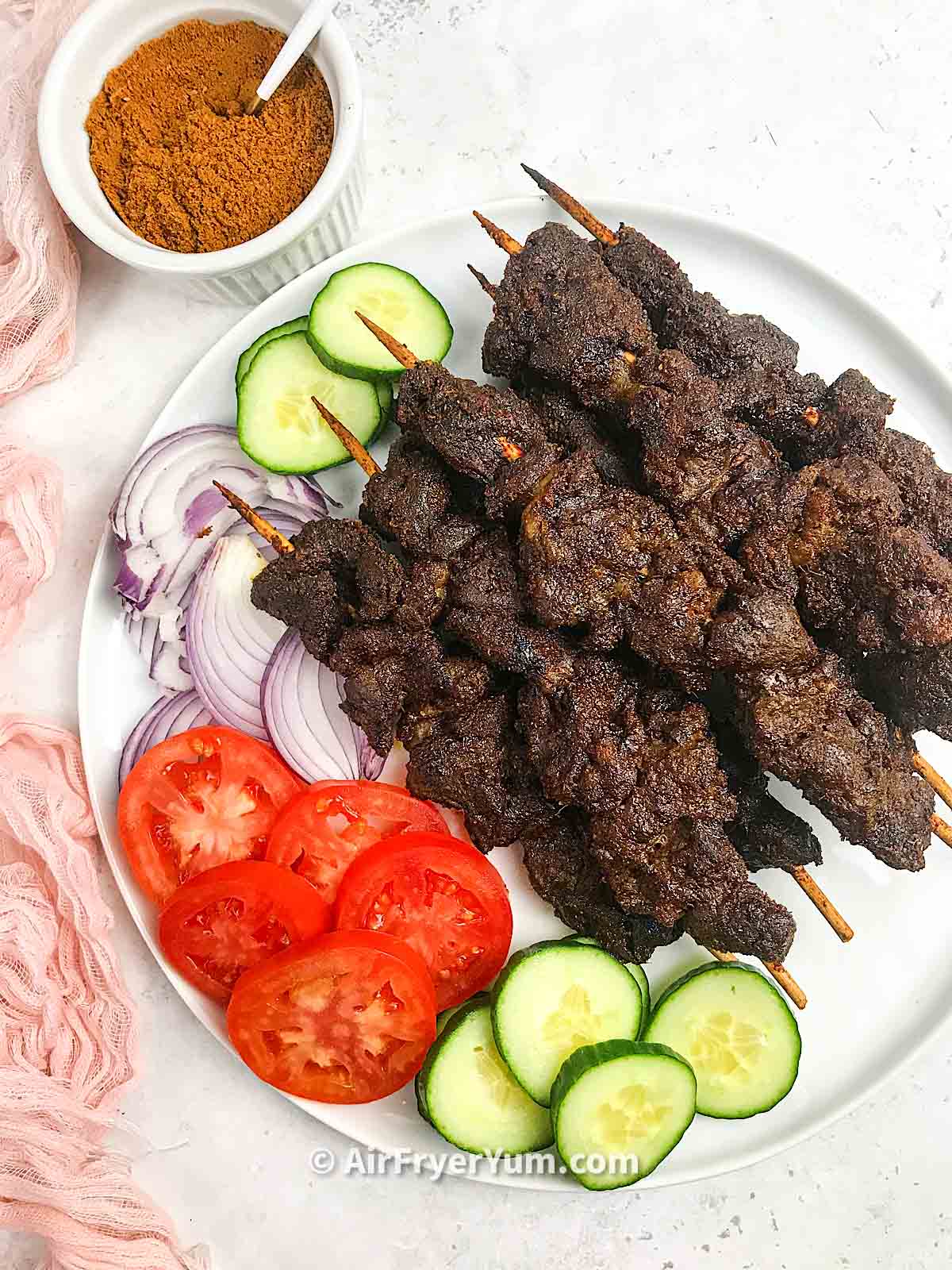 skewered beef on a white plate with some slices of tomatoes, cucumbers and onions on the side.