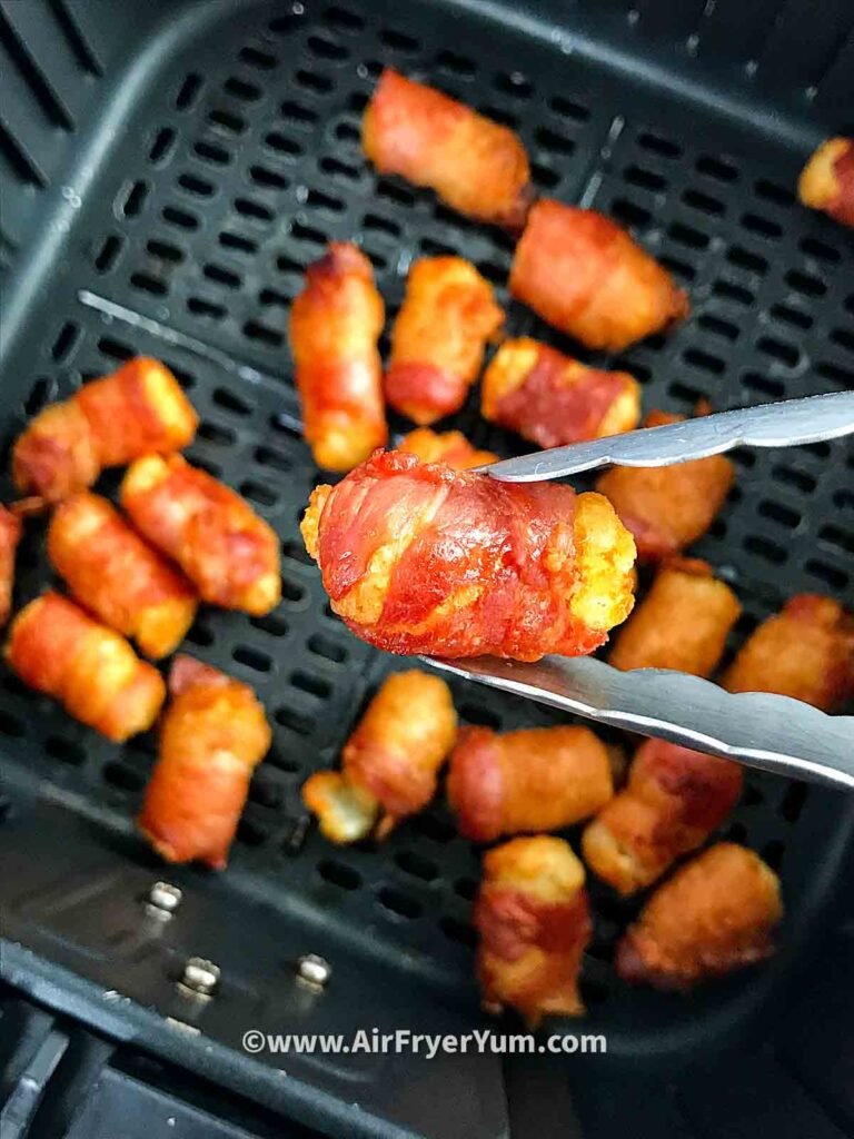 Some tater tots wrapped in bacon in an air fryer basket with one in close view on a tip of a pair of kitchen tongs 