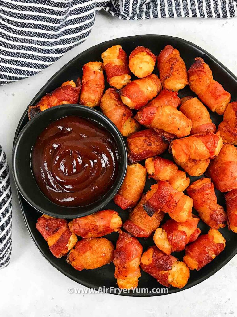 A back plate of wrapped tater tots with a black dipping bowl of bbq sauce